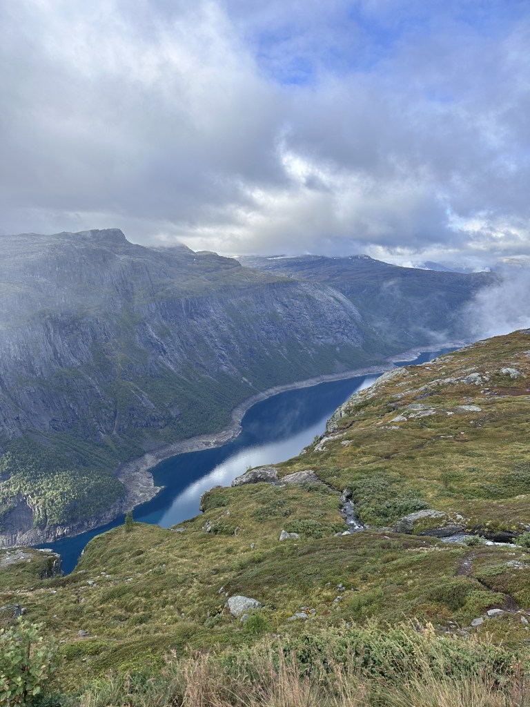 A scenic view of a valley with steep mountains and a serene lake reflecting the sky, surrounded by lush greenery and cloudy skies.
