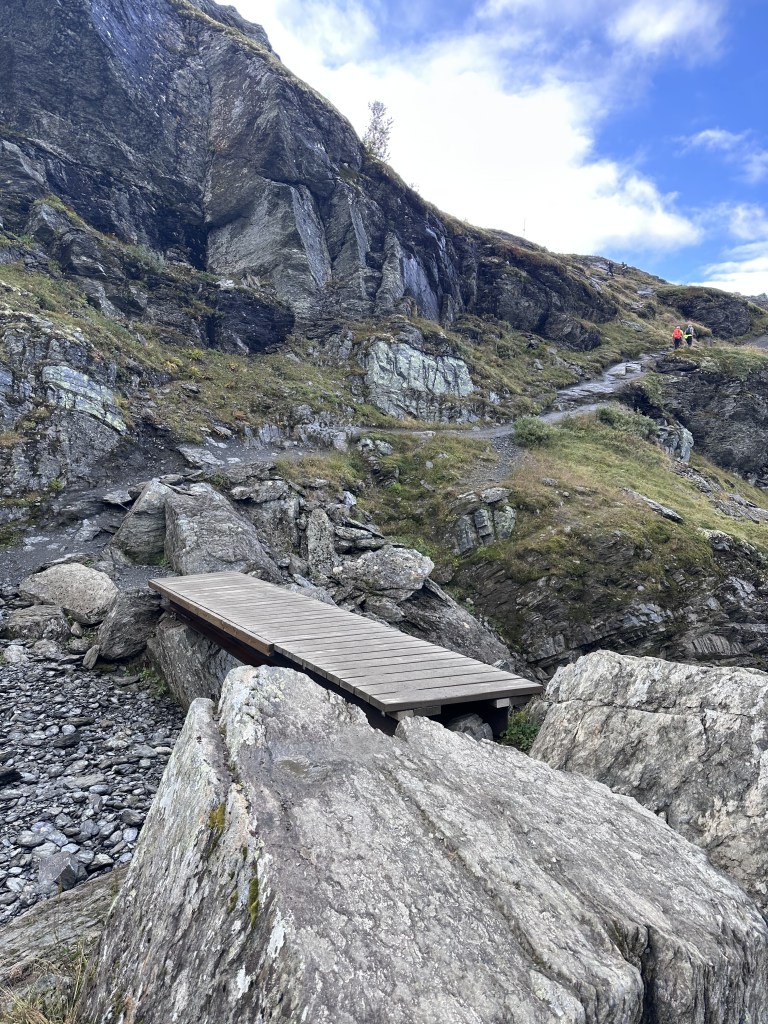 A wooden bridge spans over rocky terrain leading to a pathway on a hillside, surrounded by large boulders and steep cliffs under a blue sky.