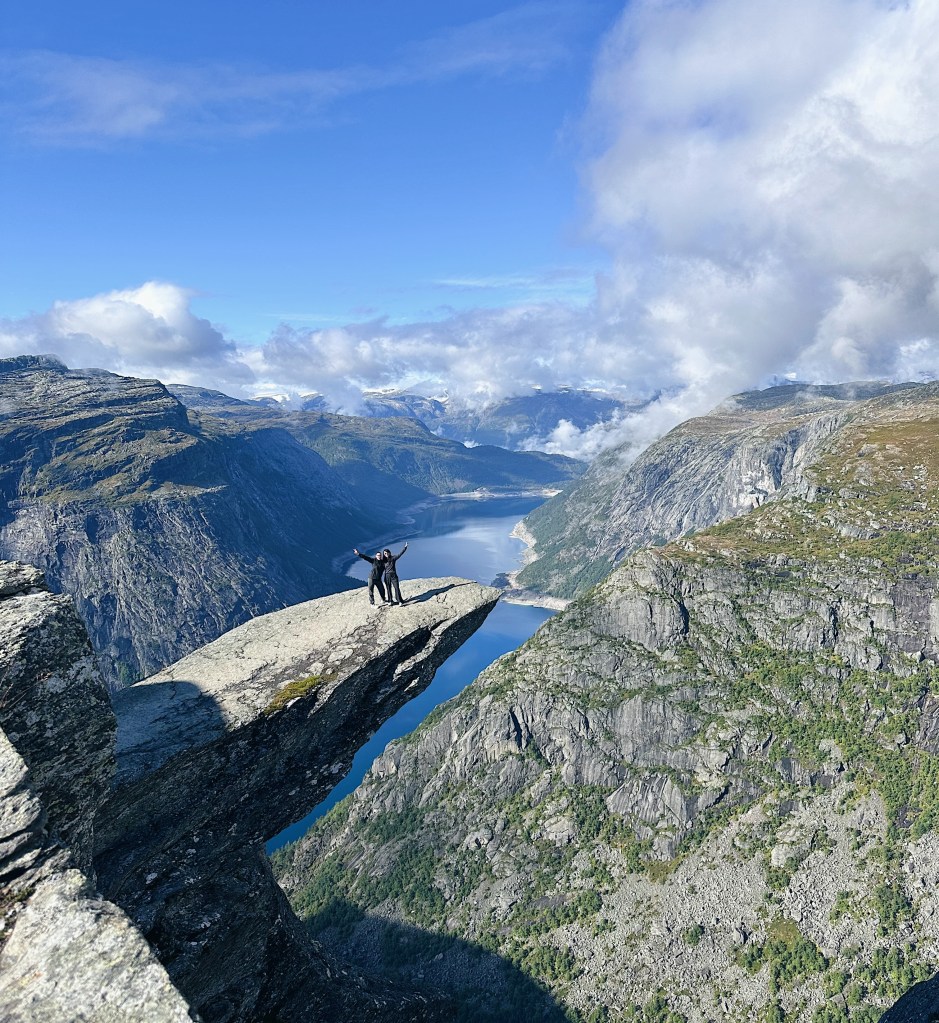 A person standing on a rocky cliff edge with arms raised in celebration, overlooking a deep fjord surrounded by mountains and a blue sky with clouds.