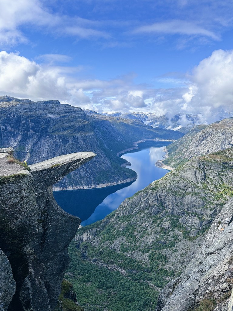 A stunning view from a rocky ledge overlooking a deep blue fjord surrounded by steep cliffs and mountains under a partly cloudy sky.
