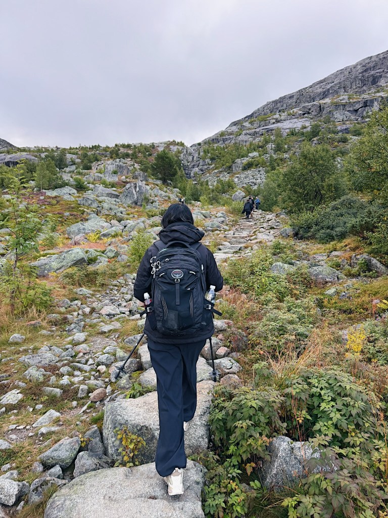 A person walking along a rocky hiking trail with a backpack, surrounded by lush greenery and steep hills under a cloudy sky.