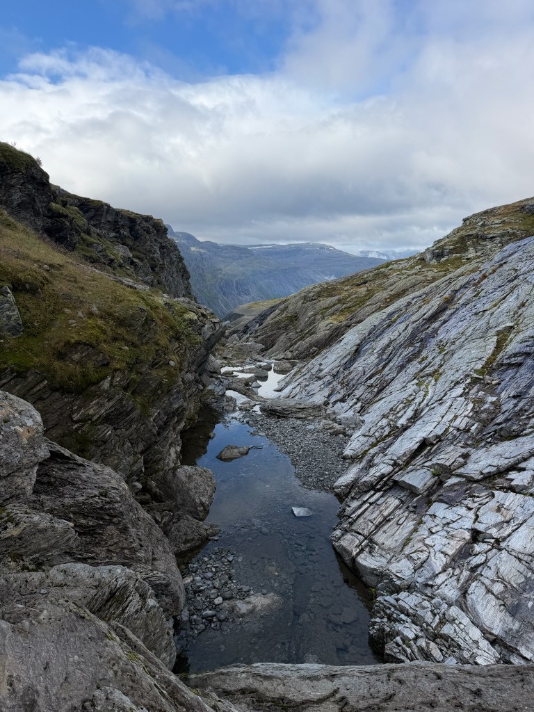 A scenic view of a rocky canyon with a shallow stream running through it, surrounded by steep rock formations and greenery, under a partly cloudy sky.