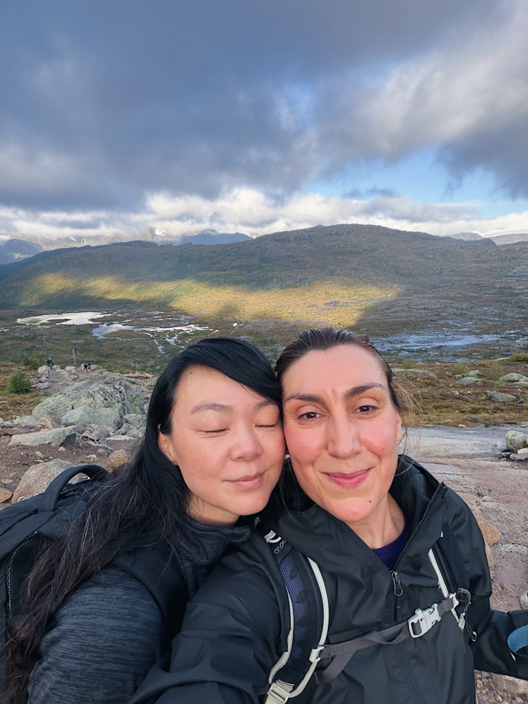 Two women smiling for a selfie in a mountainous landscape with rocky terrain and cloudy skies.