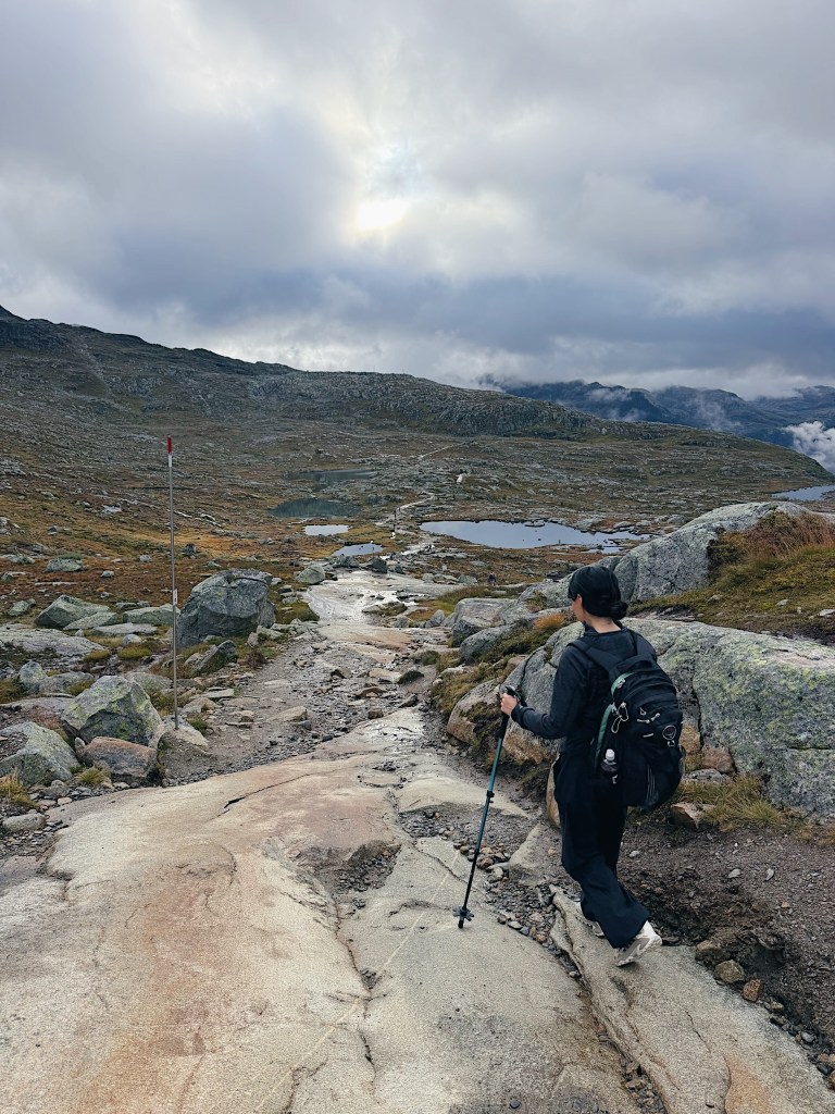 A hiker walking down a rocky path in a mountainous landscape, with cloudy skies and small ponds visible in the background.
