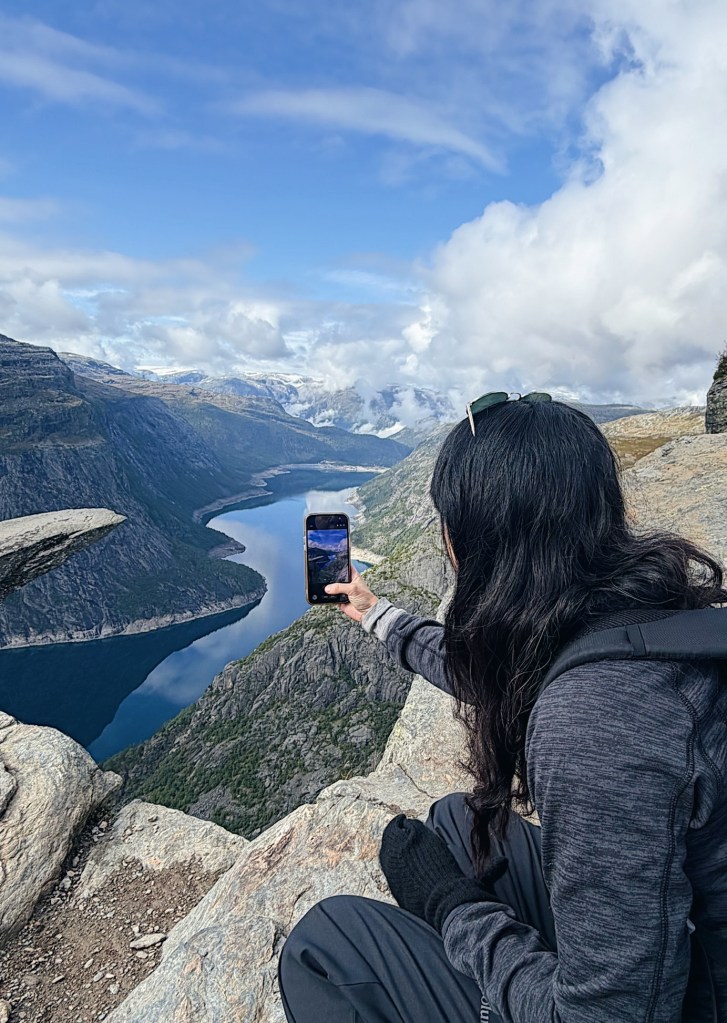 A person sitting on a rocky outcrop, holding a smartphone to capture a photo of a scenic landscape with mountains and a river below, under a partly cloudy sky.