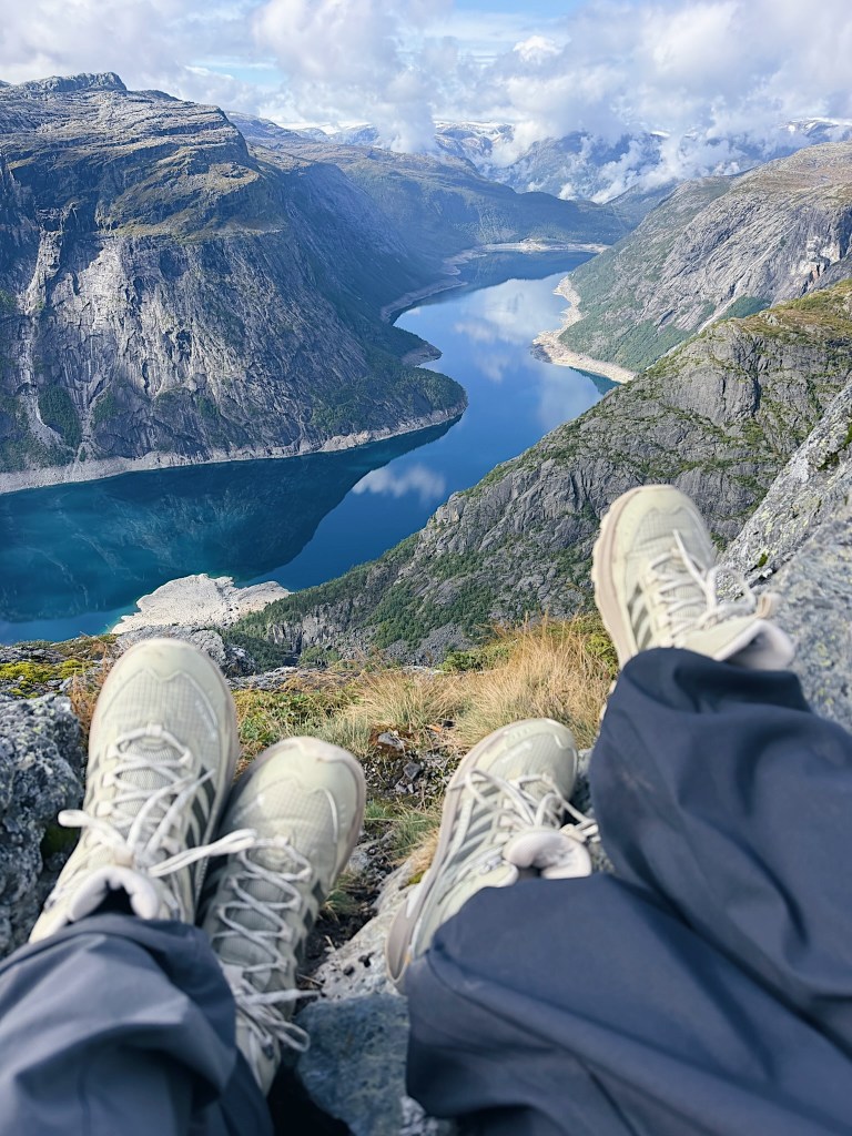 A person's feet in hiking boots resting on a rocky ledge, overlooking a scenic view of a river winding through mountains under a cloudy sky.
